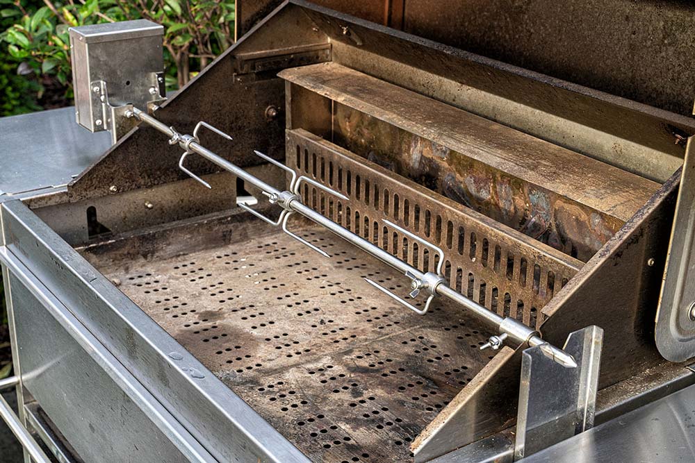 Die Grillroste sowie den Warmhalterost des Grills, in unserem Fall der Schickling-Grill Premio XL II entnehmen, den Holzkohle Backburner sowie den Rotisserie-Motor anschrauben und anschließend den Drehspieß einhängen.