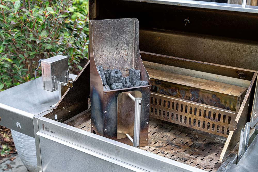 Den Backburner befüllen wir mit einem prall gefüllten Edelstahl Anzündkamin. Als Zündstoff verwenden wir die Briketts von Greek Fire.