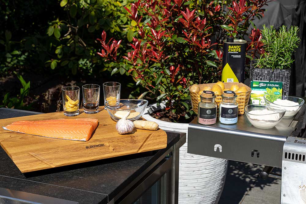 Seitenablage eines Gasgrills, daneben ein Tisch. Beides ist dekoriert mit Grillzubehör und Zutaten zum Grillen.